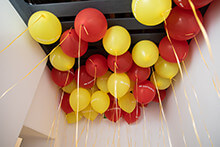 Red and yellow balloons with ribbons hanging from the ceiling in a festive indoor setting.
