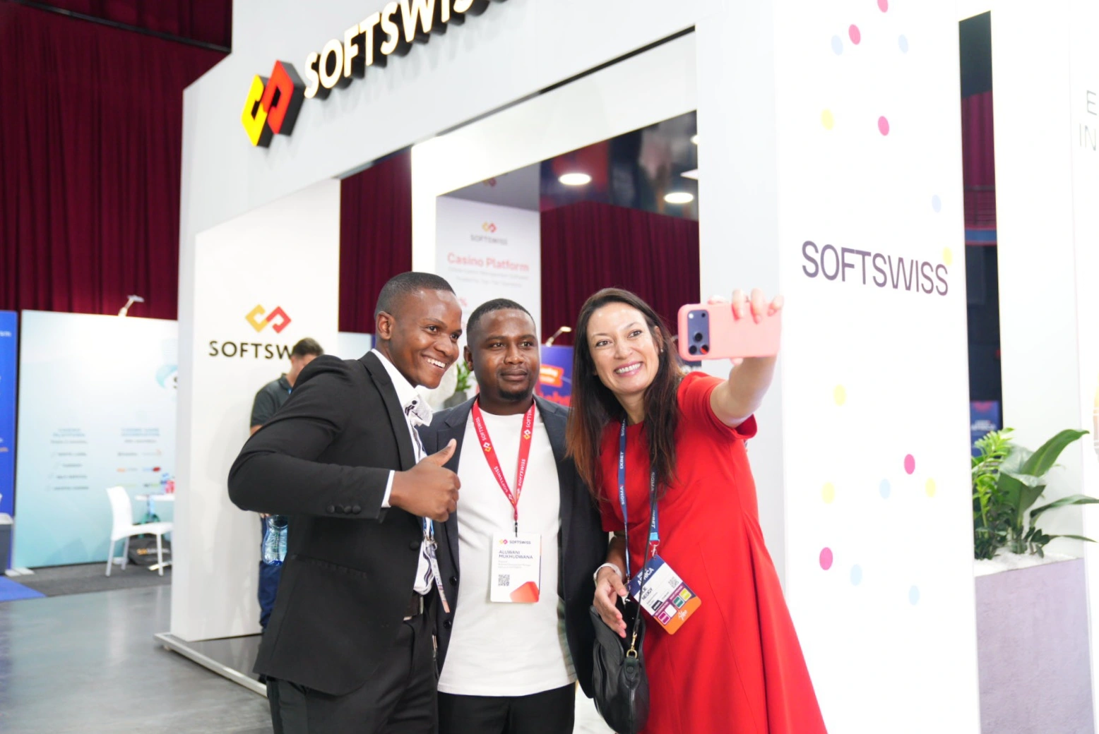 3 people taking a selfie, white SOFTSWISS booth in the background, SOFTSWISS logo